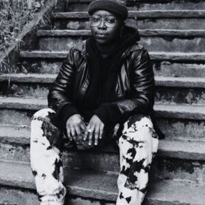 Jean poses for Trans Emergency Fund sitting on stairs and gently looking at the camera. 